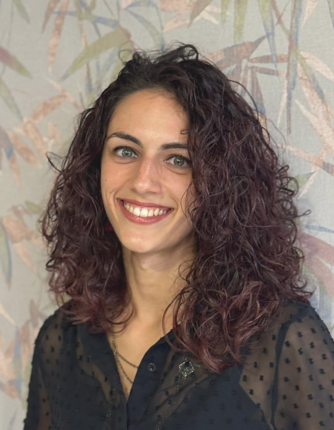 Donna con i capelli ricci, tinti di rosso, che sorride alla telecamera. Si trova in un salone con un logo AR verde.