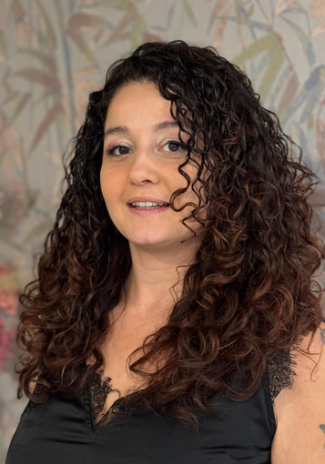 Una donna con i capelli ricci castano rossiccio sorride davanti a un cartello verde e bianco.