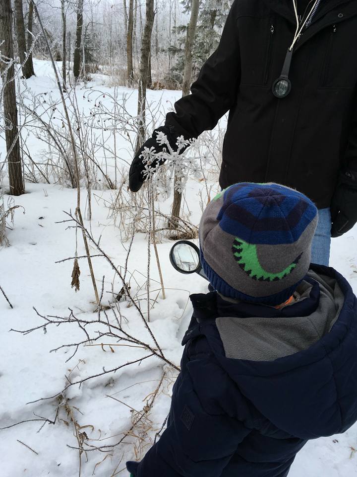 A boy wearing a dinosaur hat is looking through a magnifying glass in the snow.