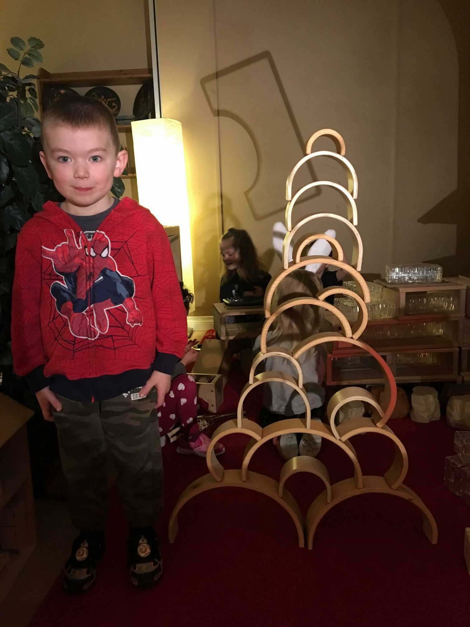 A young boy wearing a spiderman shirt is standing next to a stack of wooden arches