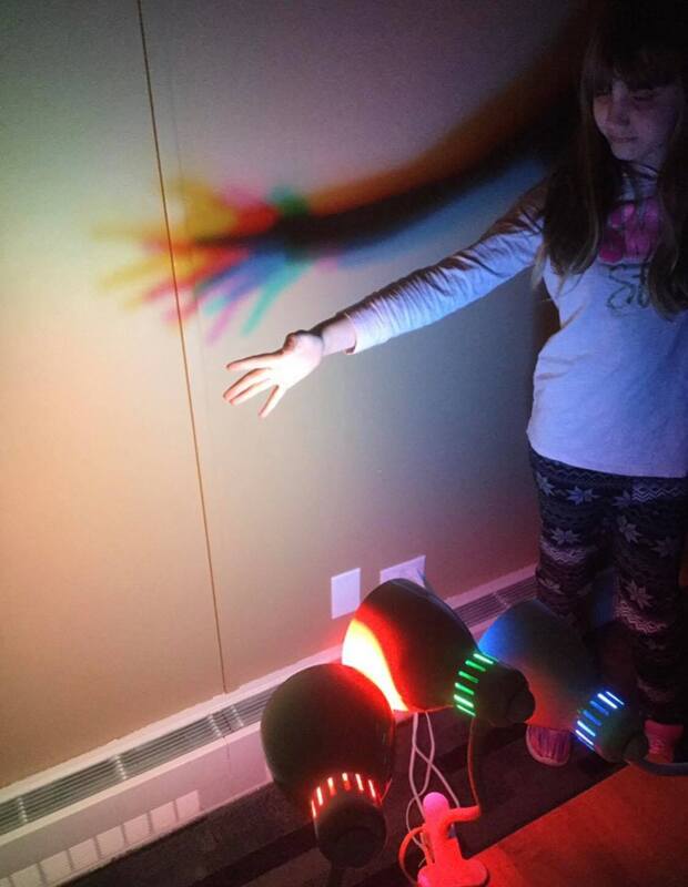 A little girl is standing in front of a wall with a rainbow projected on it