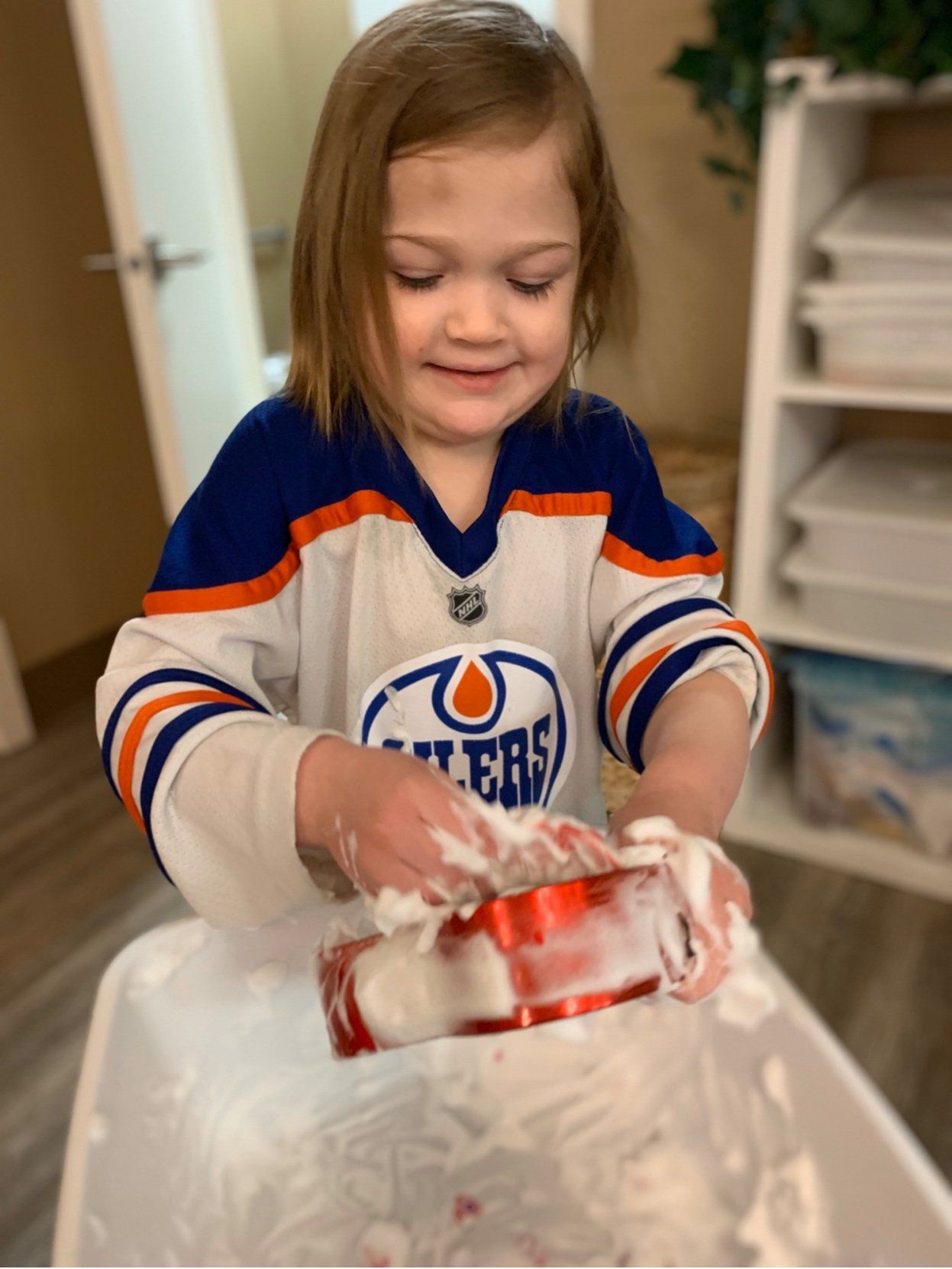 A little girl in an edmonton oilers jersey is playing with shaving cream.
