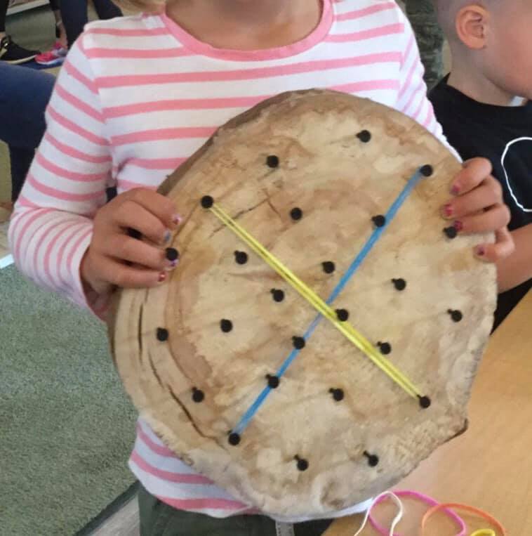 A girl in a pink and white striped shirt is holding a piece of wood with holes in it