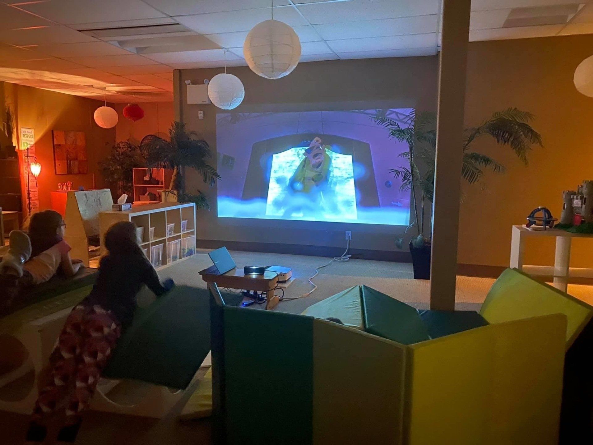 A group of people are sitting in a living room watching a movie on a projector screen.