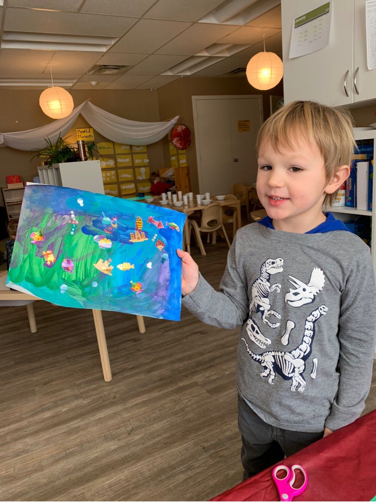 A young boy is holding up a painting in a classroom.