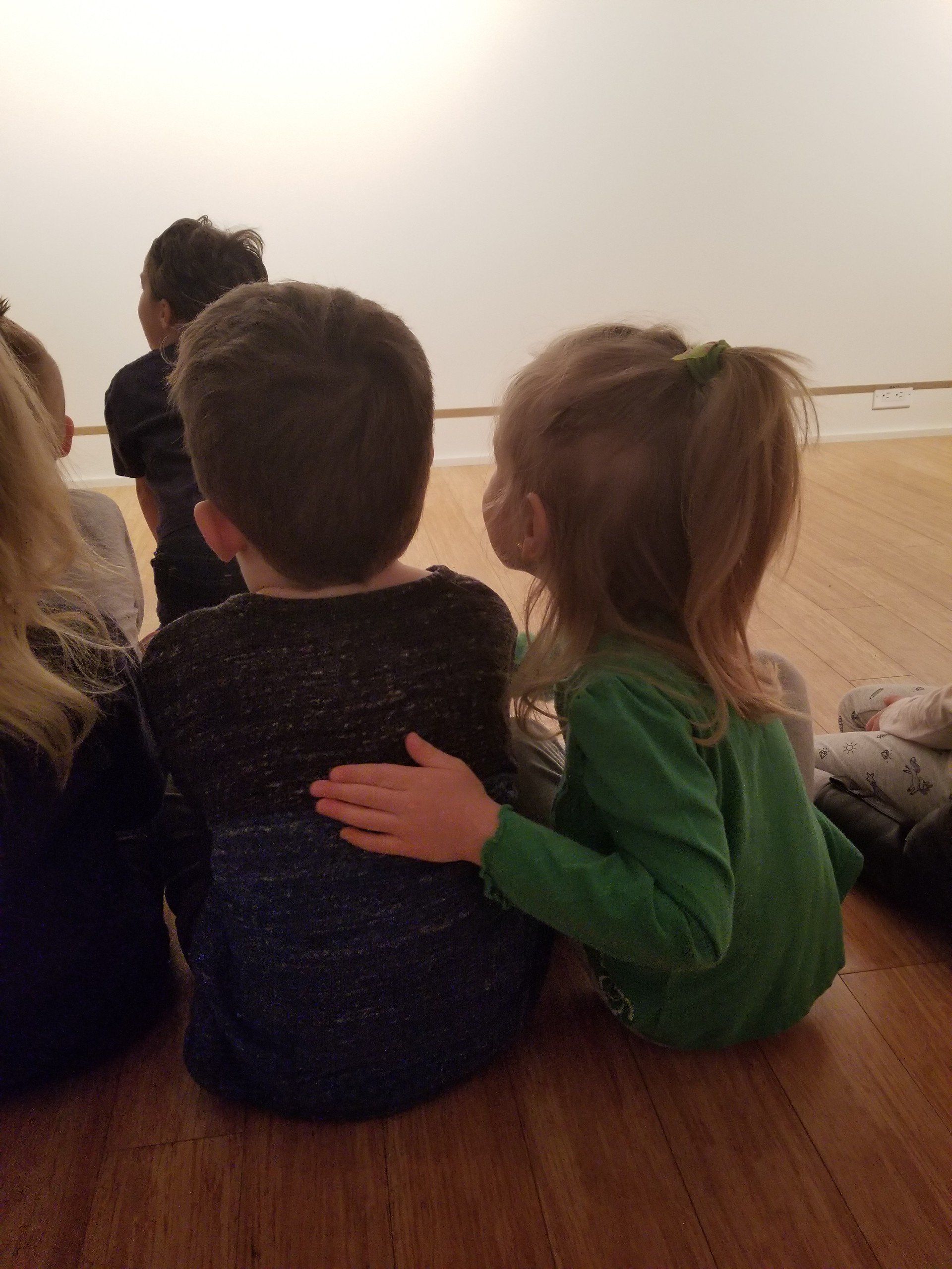 A group of children are sitting on the floor looking at a white wall.