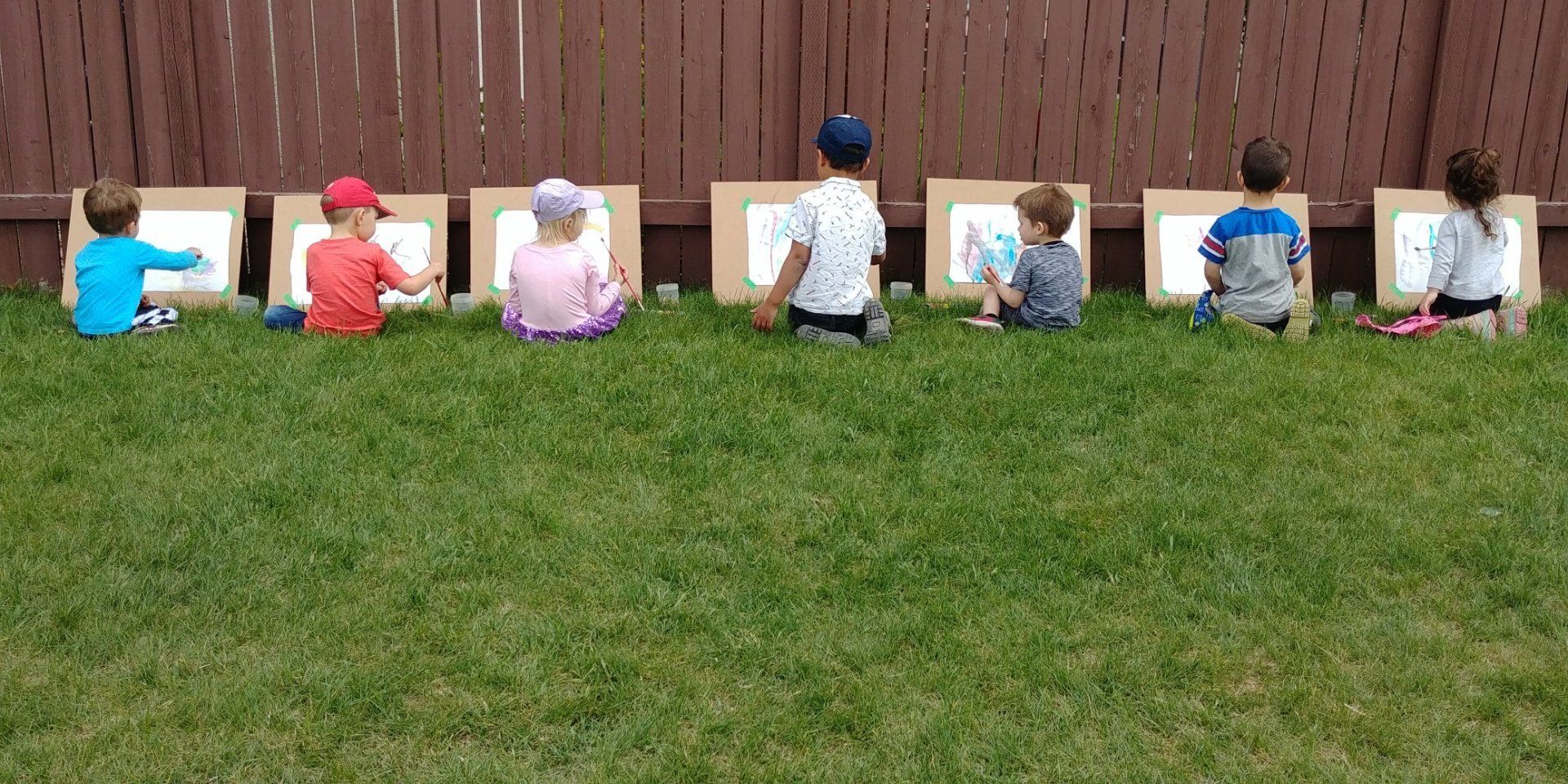 A group of children are sitting in the grass with cardboard boxes.