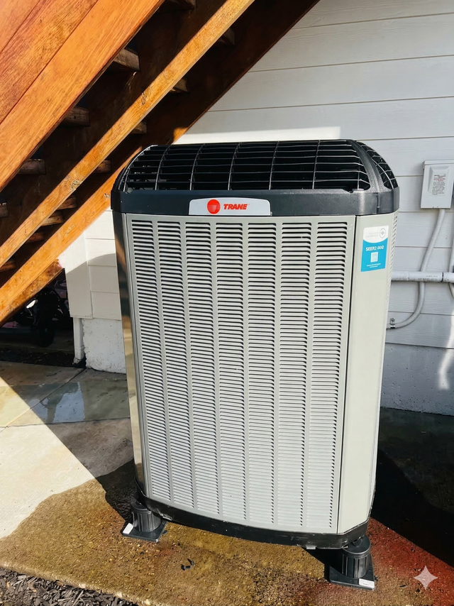 A Trane air conditioning unit sits on a concrete pad outdoors beneath a wooden staircase.