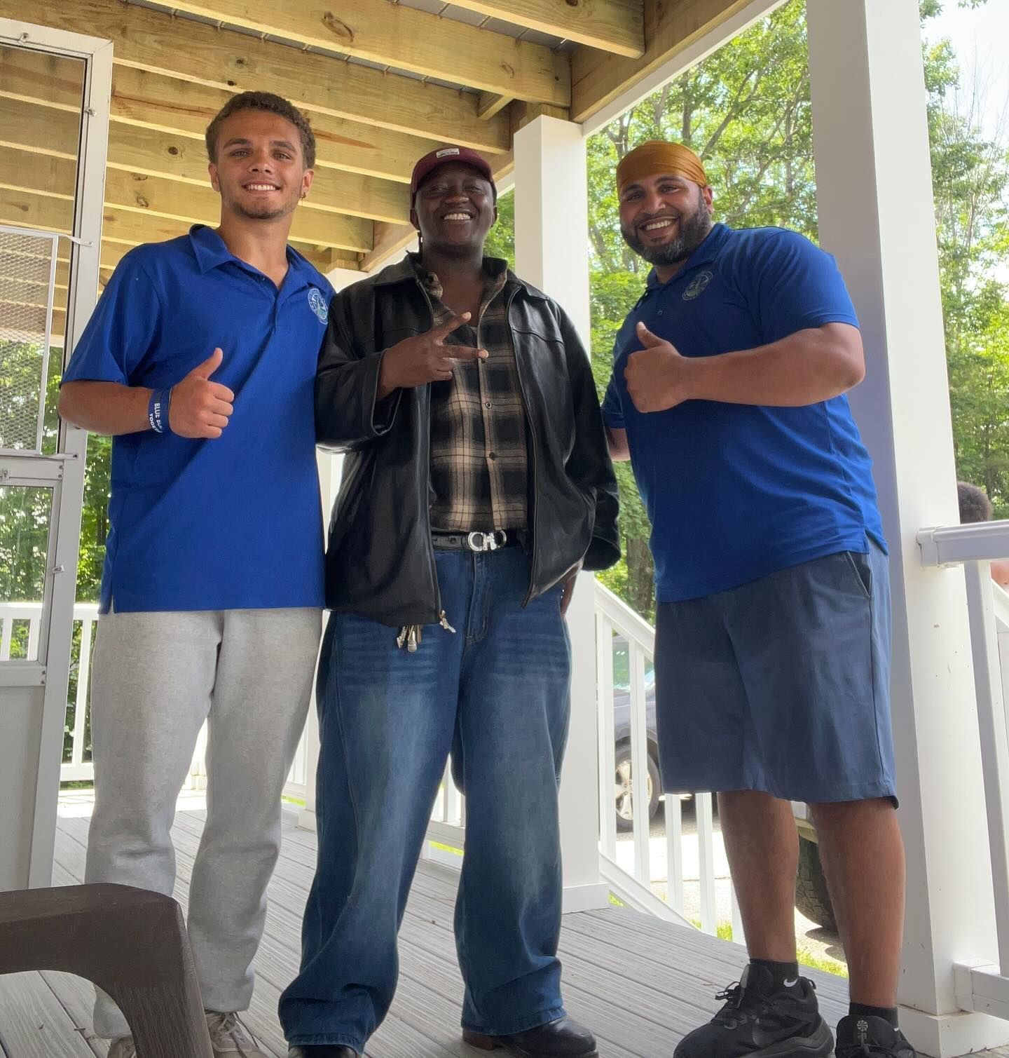 Three people smiling on a porch. Middle person in leather jacket, denim jeans. Others in blue shirts, giving thumbs up.