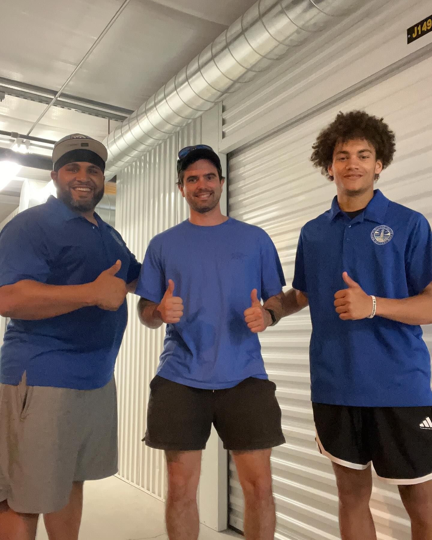 Three people in blue shirts, giving thumbs up inside a storage unit.
