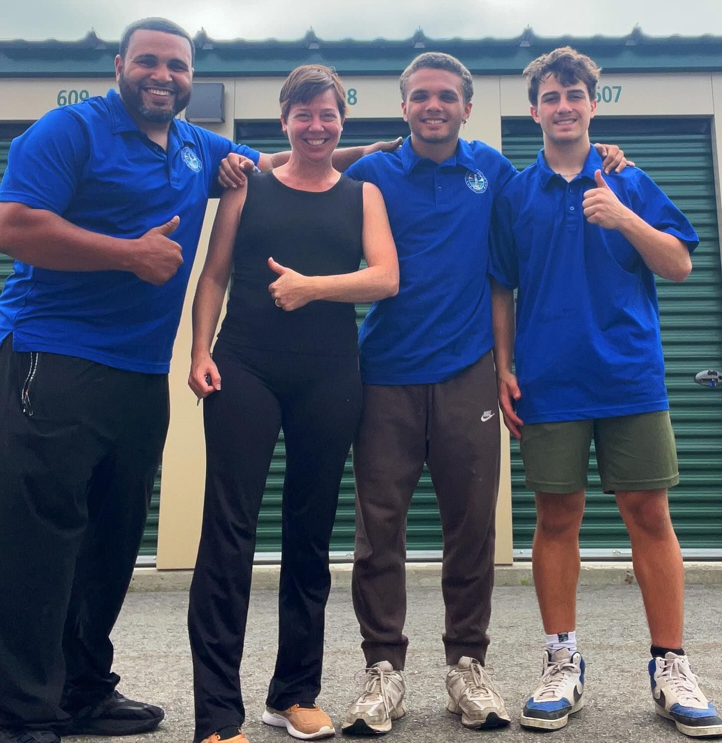 Four people in blue shirts, giving thumbs up, in front of storage units.