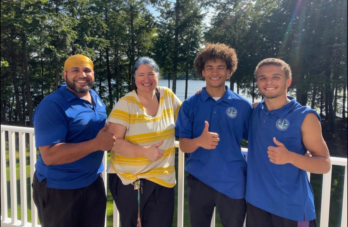Four people on a deck; two giving thumbs-up. Blue and yellow shirts; trees and water in the background.