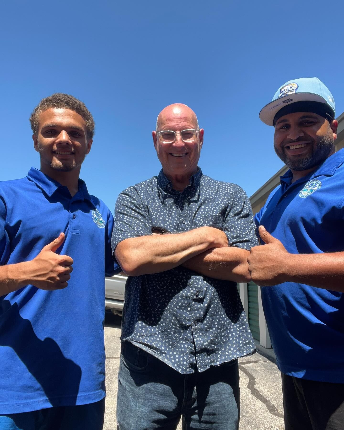 Three men smiling outdoors on a sunny day. Two in blue shirts give thumbs up, while a man in a patterned shirt has arms crossed.