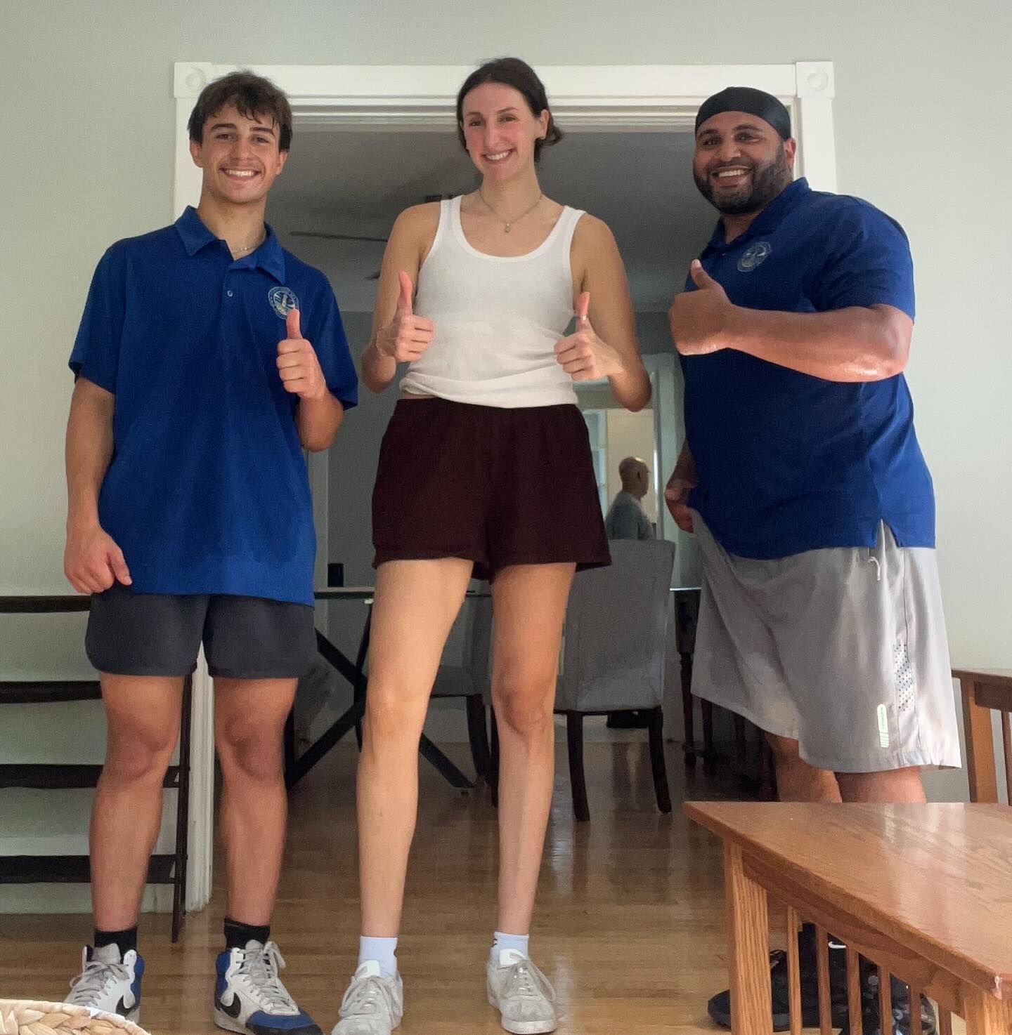 Three people giving thumbs-up inside a room. Tall woman in shorts and tank top, two men in shirts and shorts.