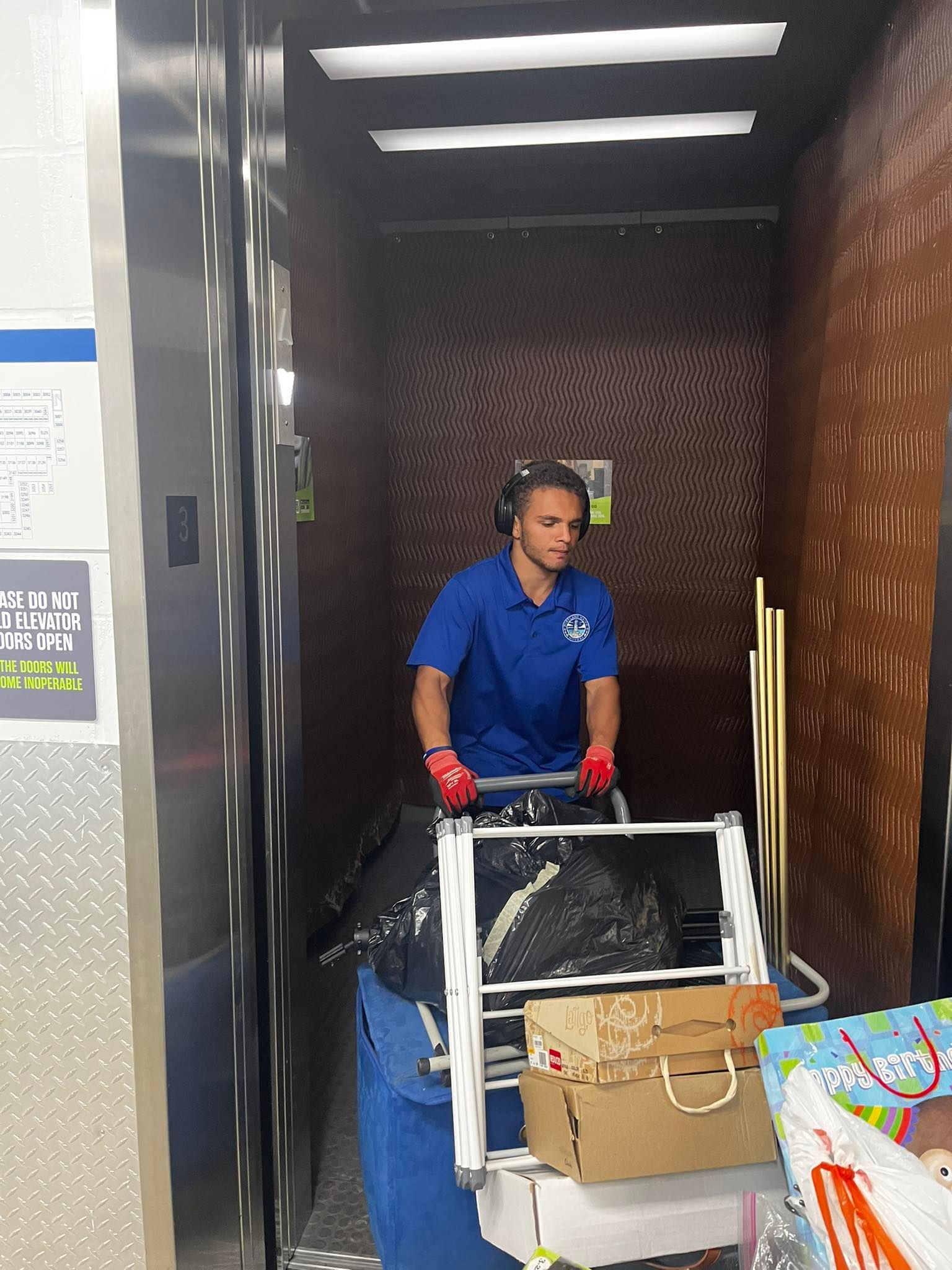 A person in a blue shirt with a cart in an elevator. The cart has bags and boxes.