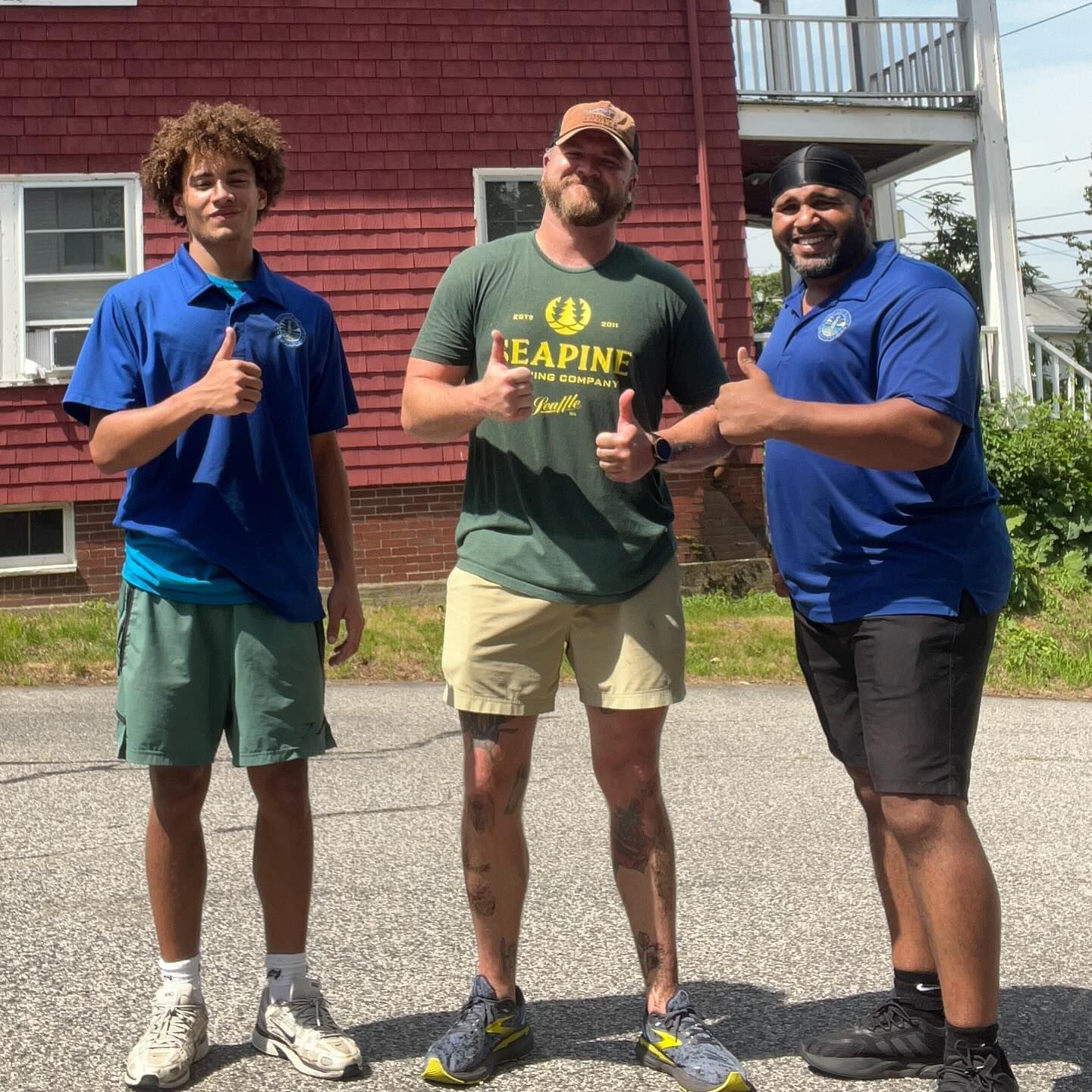 Three people give thumbs up in front of a red building; two wear blue shirts and one wears green.