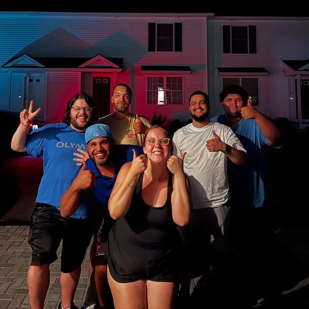Group of seven people smiling and giving thumbs up in front of a house at night, lit with blue and red lights.