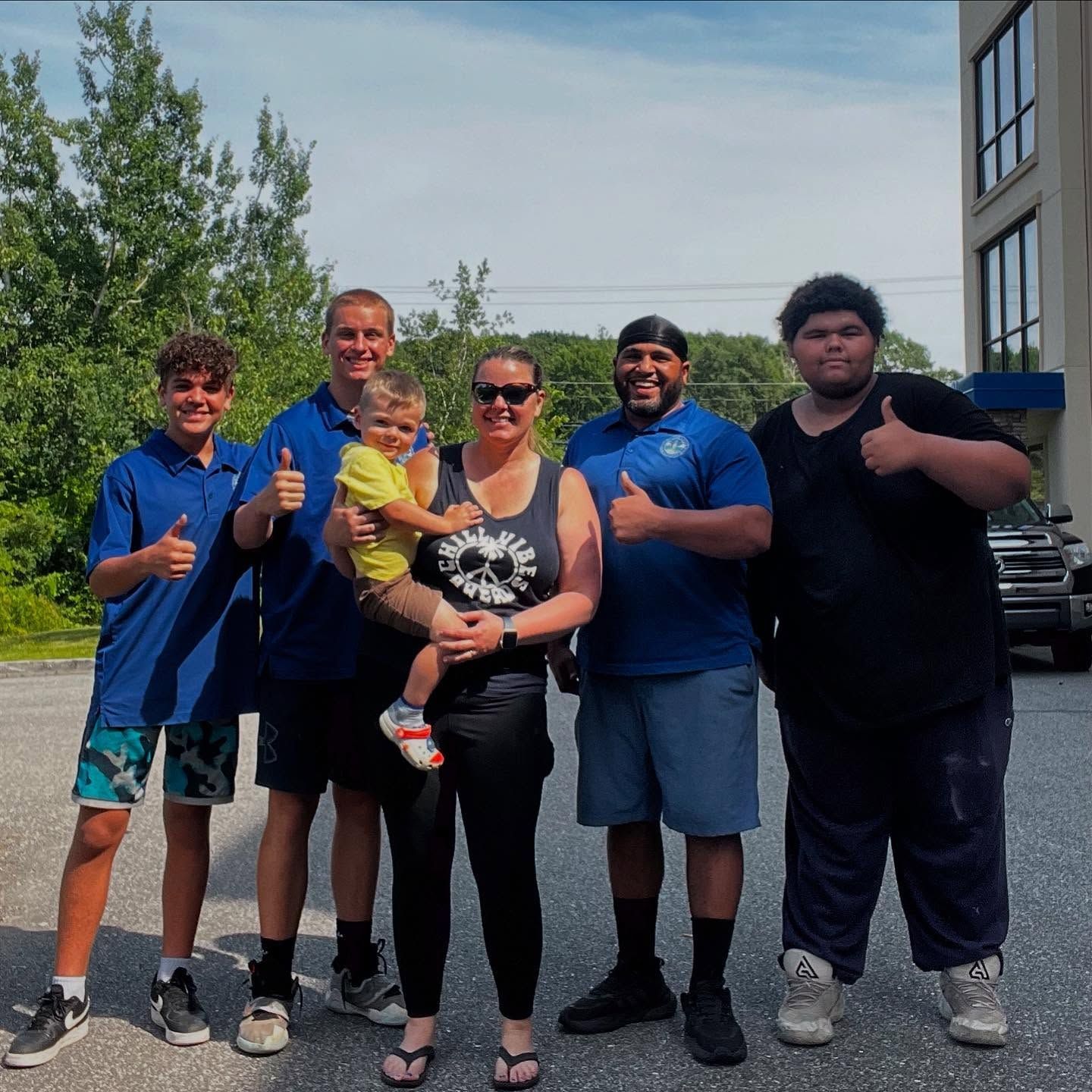 Group of people smiling and giving thumbs up outside a building. Sunny day.