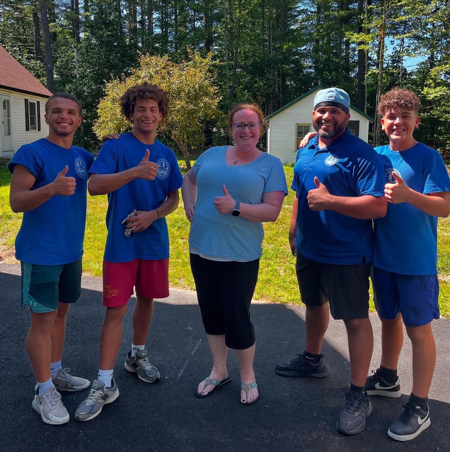 Five people in blue shirts giving thumbs up outside a building on a sunny day.