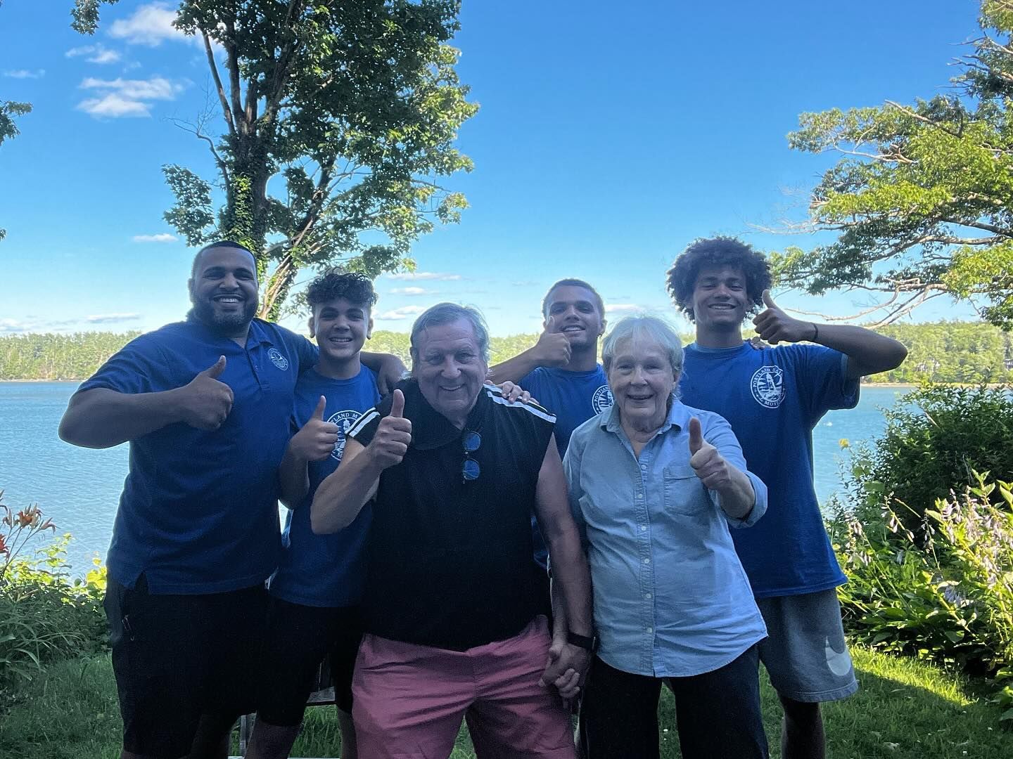 Group of six people by a lake, giving thumbs up. Blue shirts, smiling, sunny day.