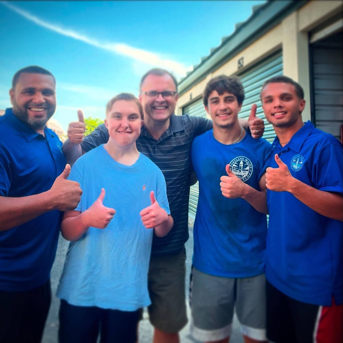Five people smiling and giving thumbs up in front of storage units. Blue shirts and sunny day.