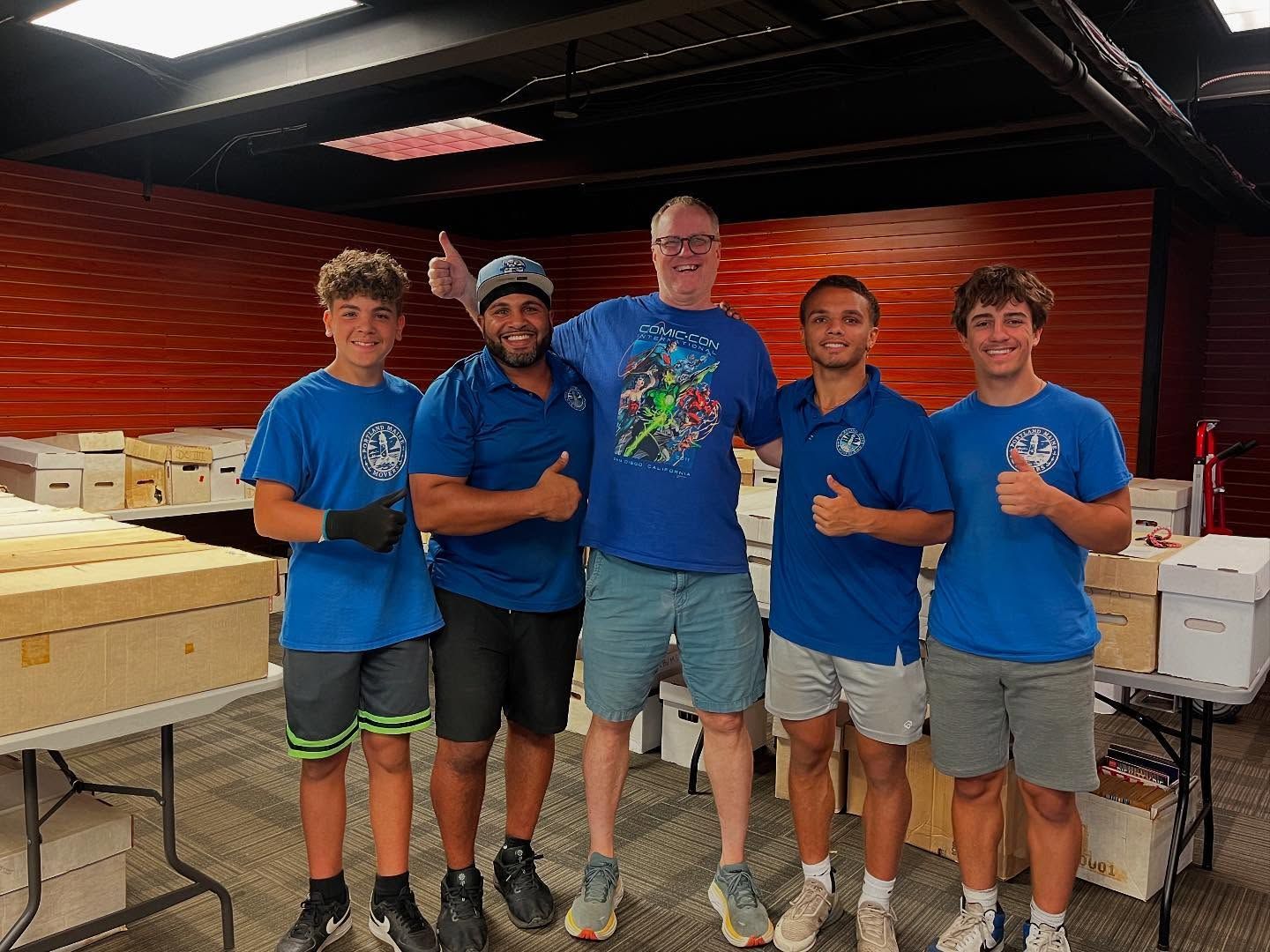 Five people in blue shirts giving thumbs up. Boxes and tables in a room with orange walls.