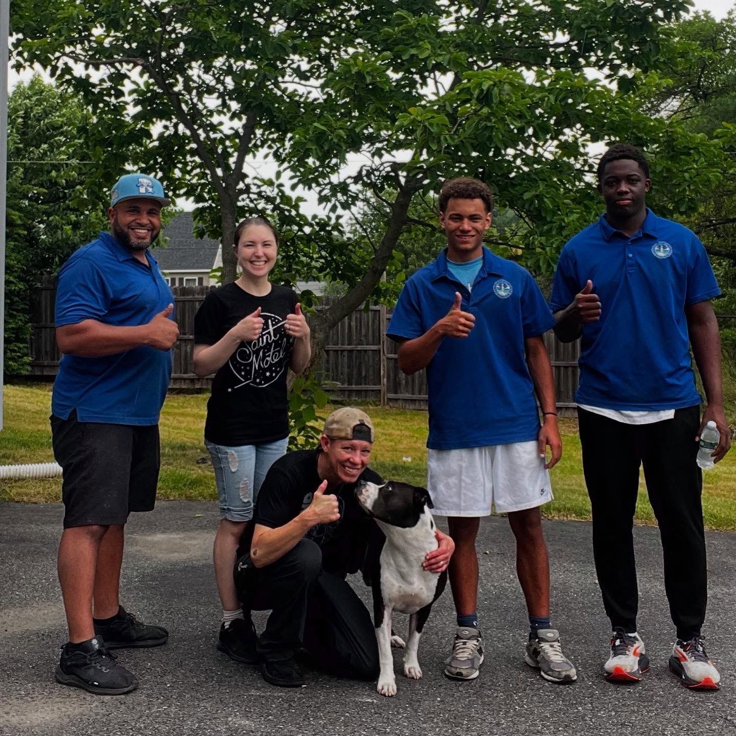 Group of people with thumbs up, smiling next to a dog outside.