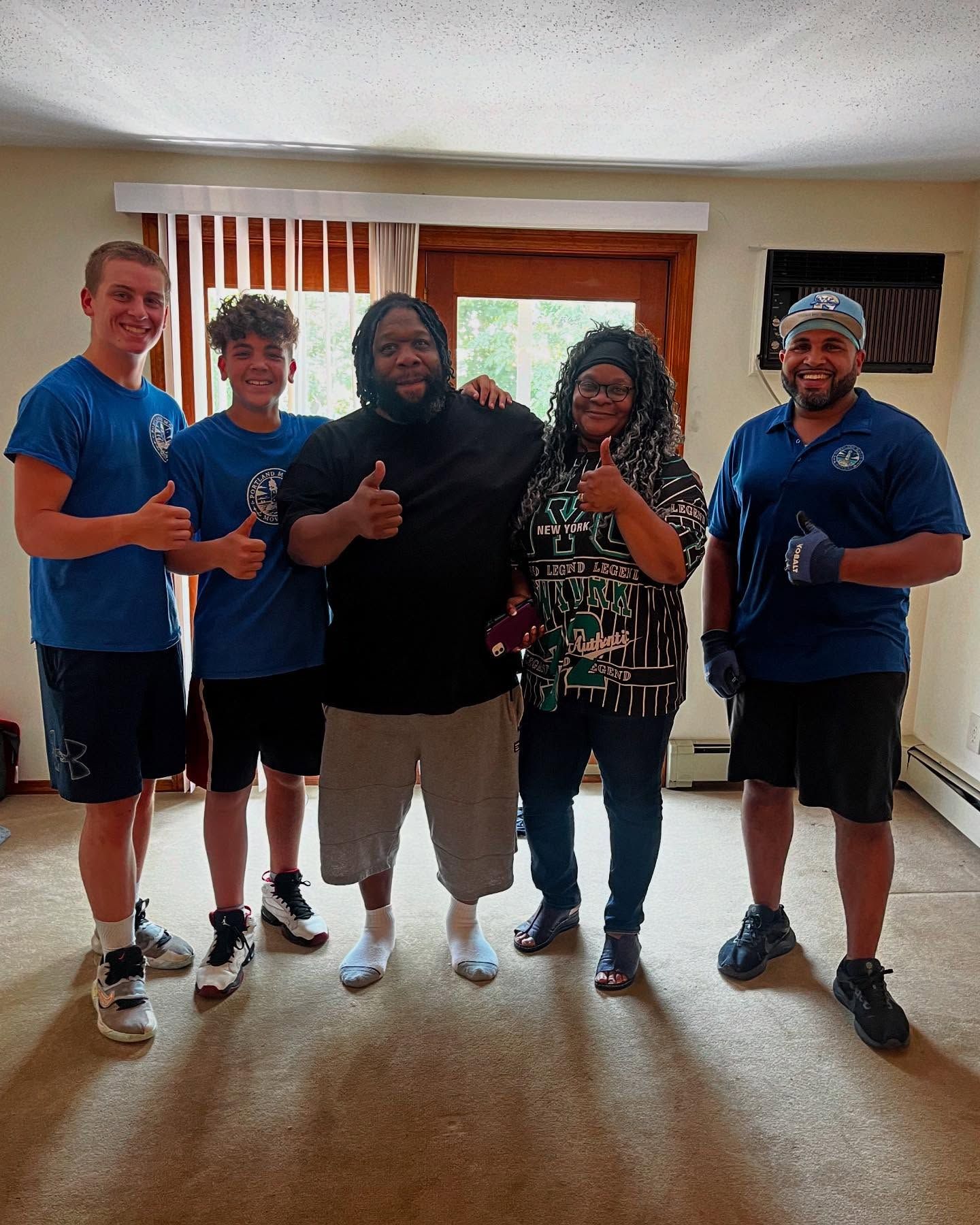 Five people giving thumbs up in an empty room with a window and door. They appear happy and satisfied.