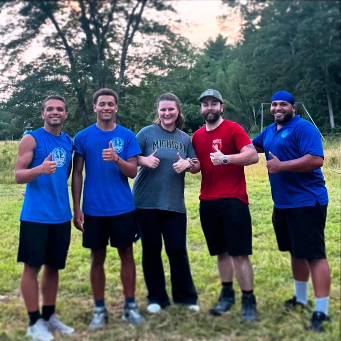 Five people giving thumbs up in a grassy field. Trees in the background, some wearing blue and red shirts.