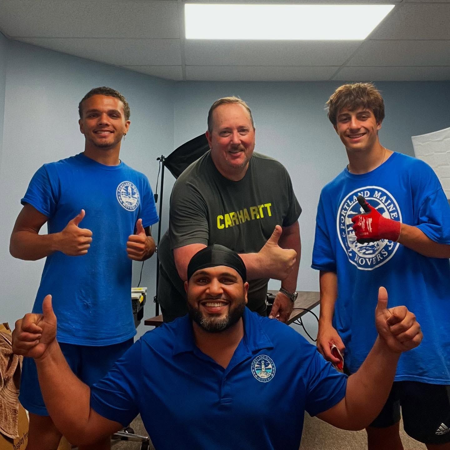 Four men in blue shirts give thumbs up. One man wears a head wrap. Indoors, neutral walls.