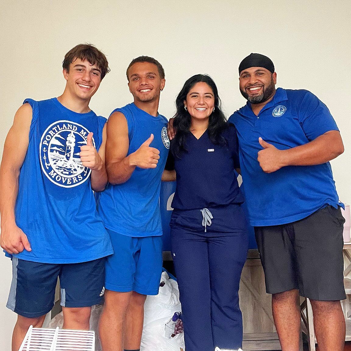 Four people in blue, giving thumbs up. Two wear blue shirts, one in scrubs, one in a cap, outdoors.
