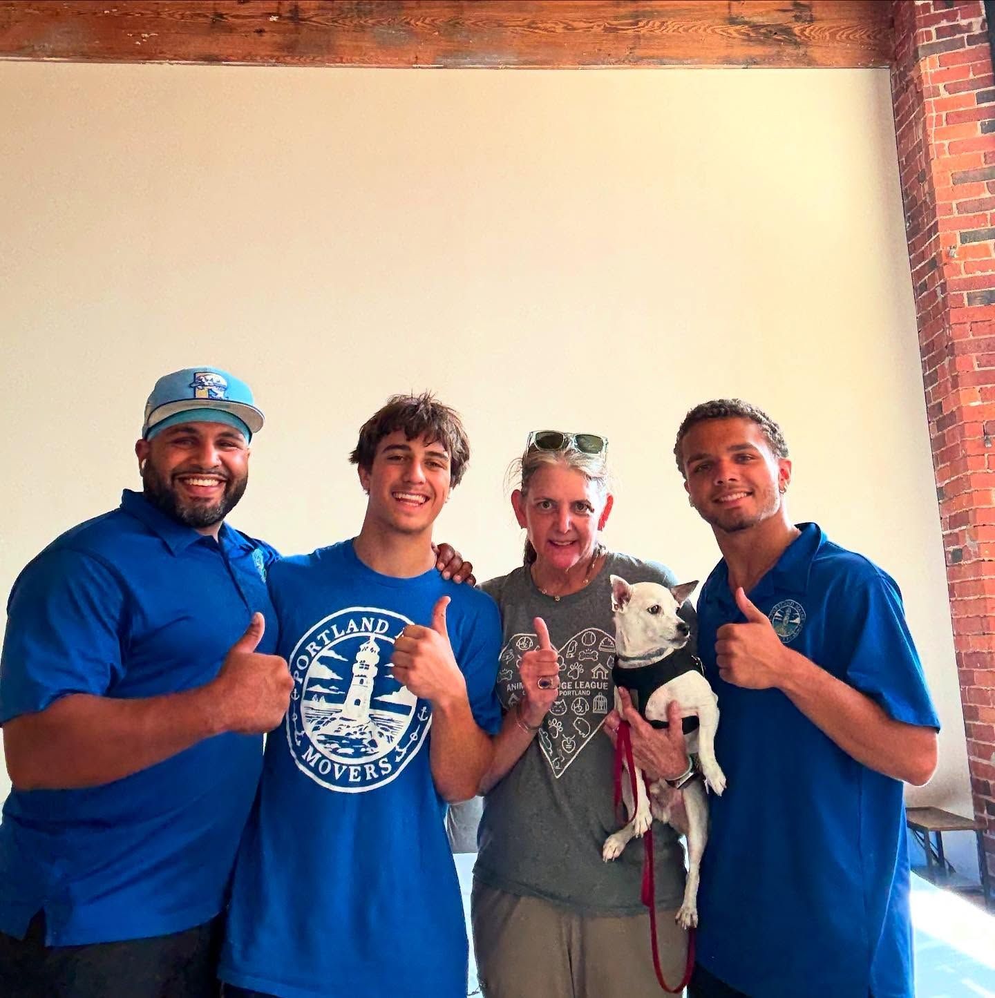 Four people and a dog give thumbs up; two men wear blue, woman holds dog, background is white and brick.