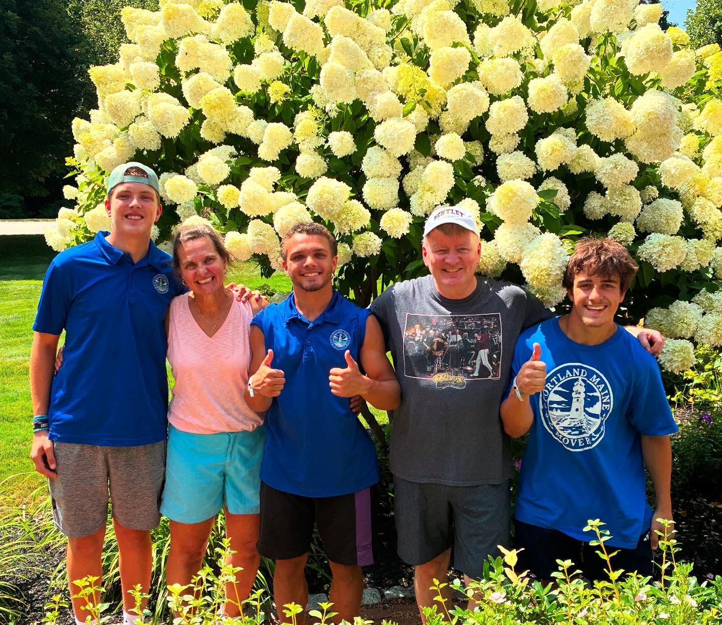 Five people smiling, giving thumbs up, in front of white flowers. Outdoors, sunny day, blue and light colors.