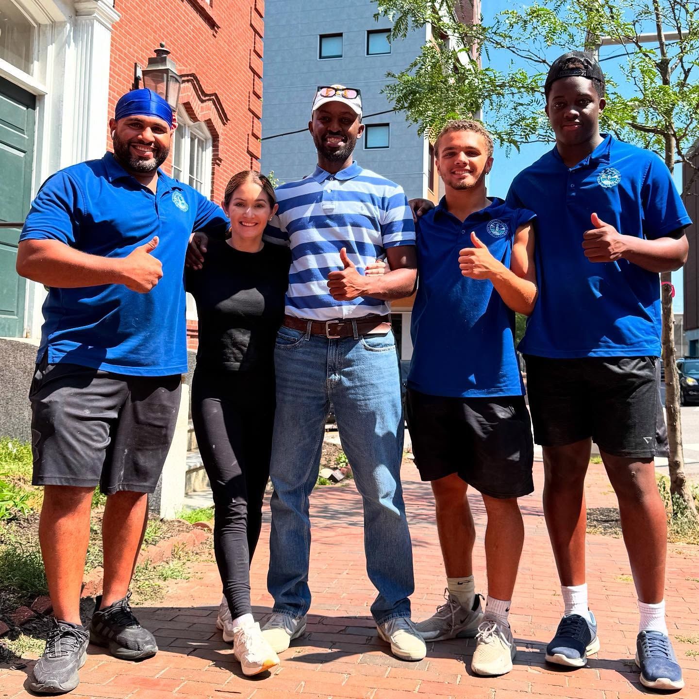 Five people smiling, giving thumbs-up. They stand on a sidewalk, outside a brick building.