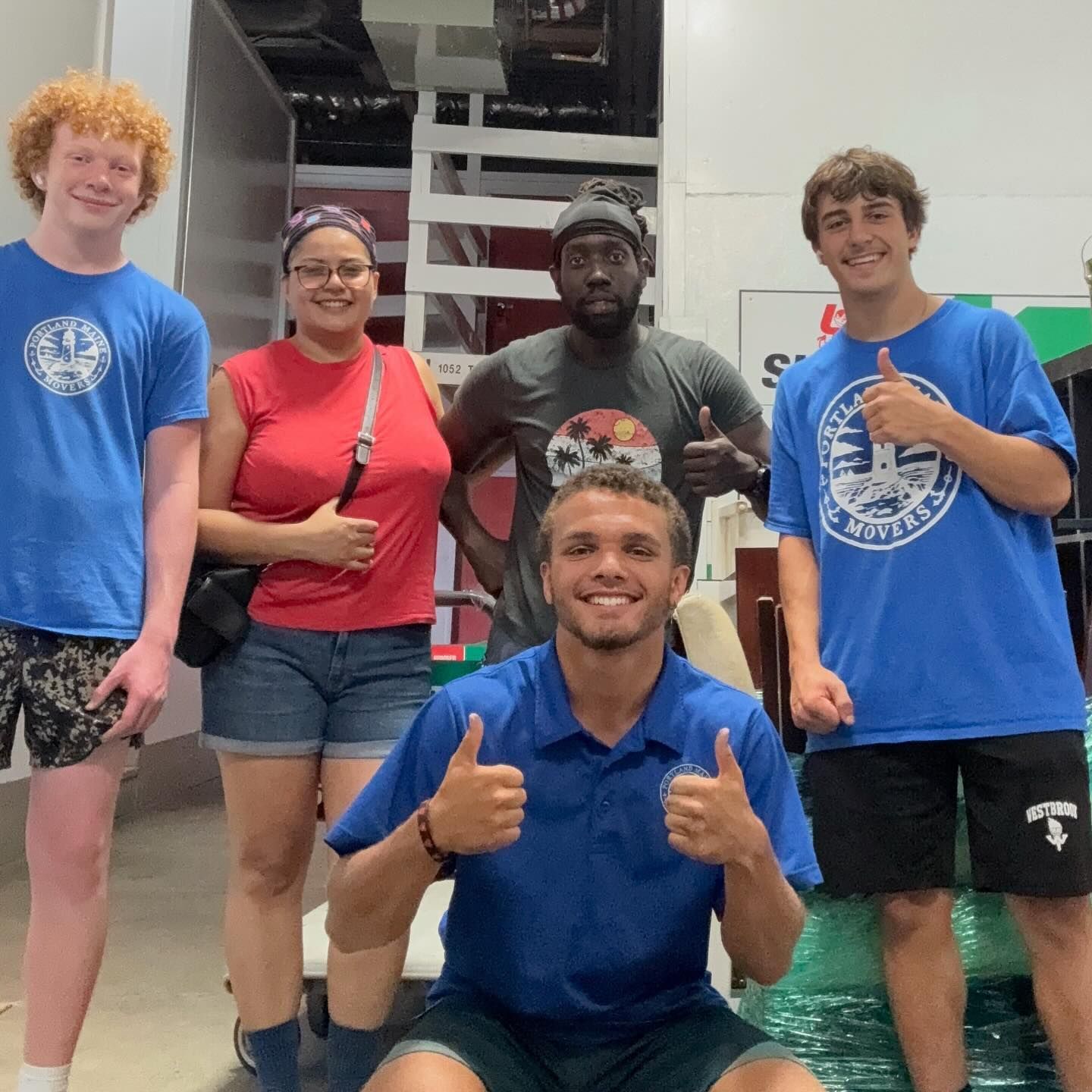 Five people, thumbs up. Inside, likely a storage facility or moving space. Smiling faces, various casual attire.