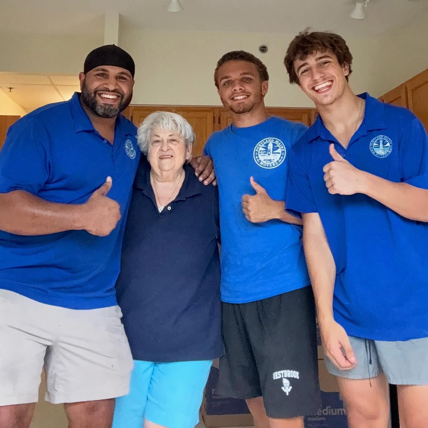 Four people, two men and a woman pose with thumbs up. They are wearing blue shirts, the men with tan shorts and the woman with blue pants.