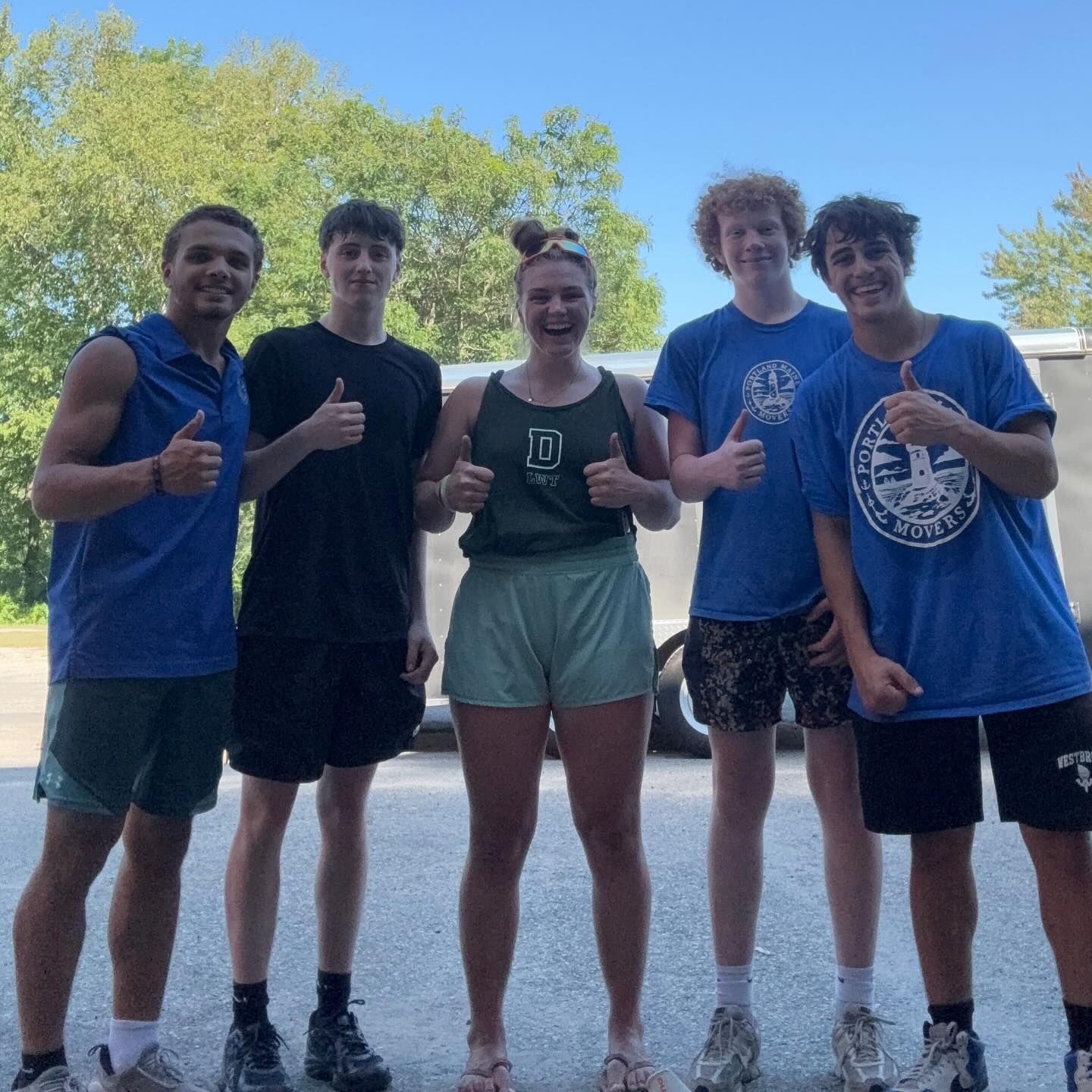 Five people smiling, giving thumbs up. Outdoors, sunny.