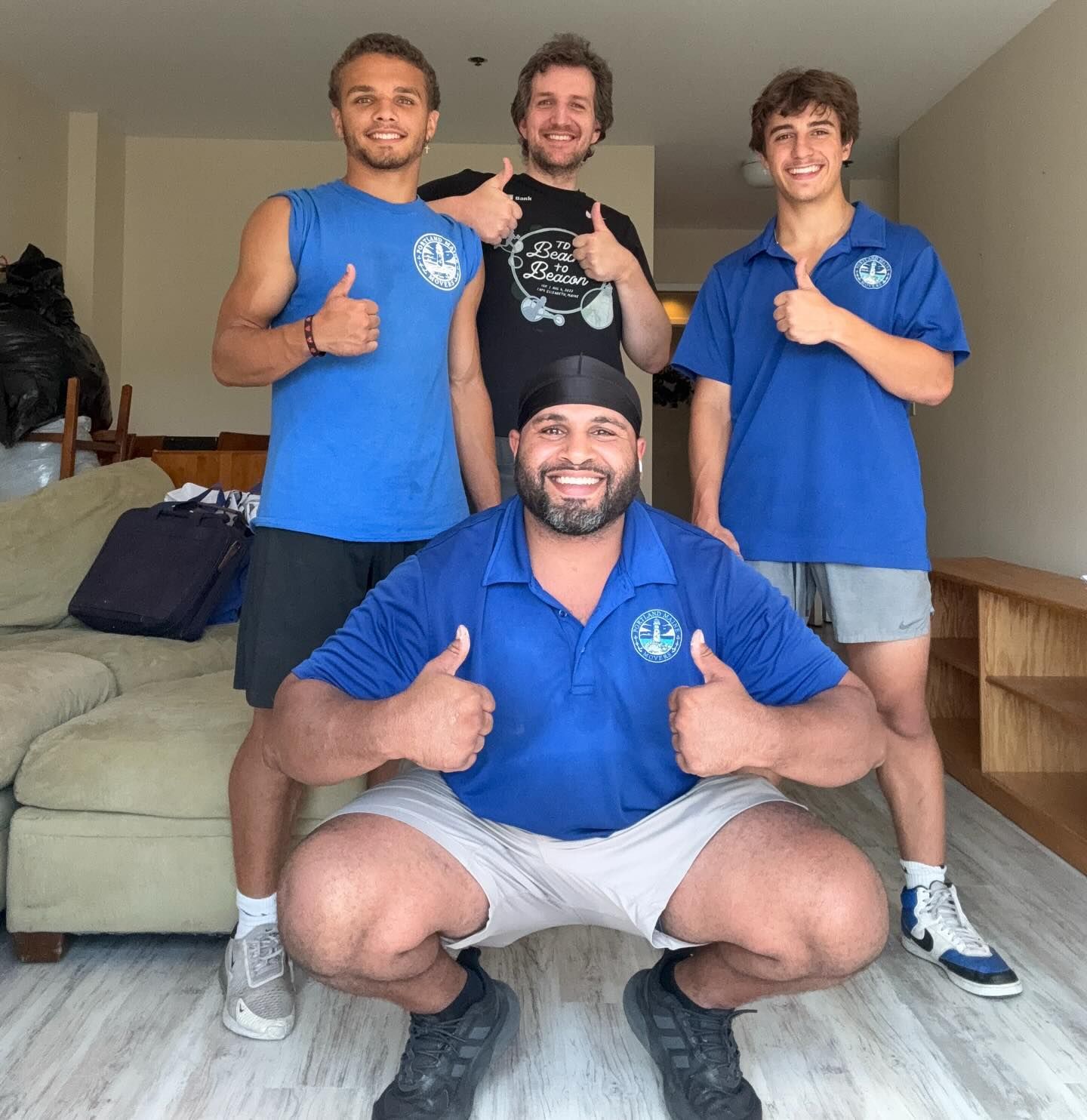 Four people, one crouching, give thumbs up. They are in a room with a couch and shelves.