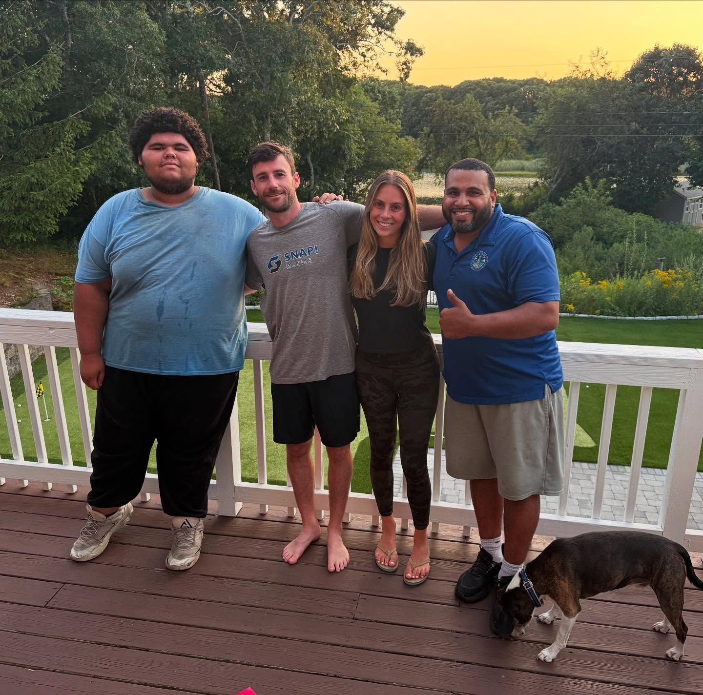 Four people pose together on a wooden deck; one gives a thumbs-up. A dog stands nearby. The backdrop is trees and a sunset.