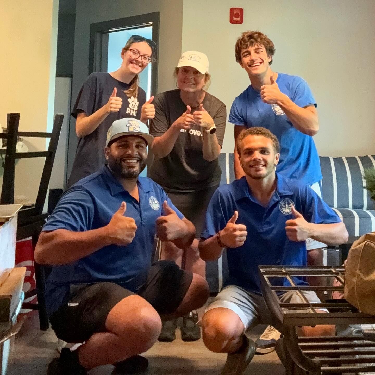 Group of people giving thumbs up in a room. Some wear blue shirts, and one has a white hat.