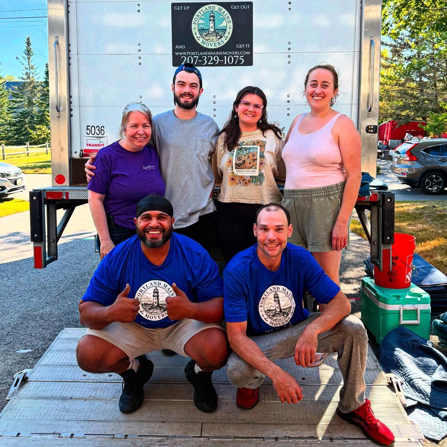 Movers posing in front of a truck, featuring a lighthouse logo. Two are squatting, others standing, smiling. Blue shirts, outside.