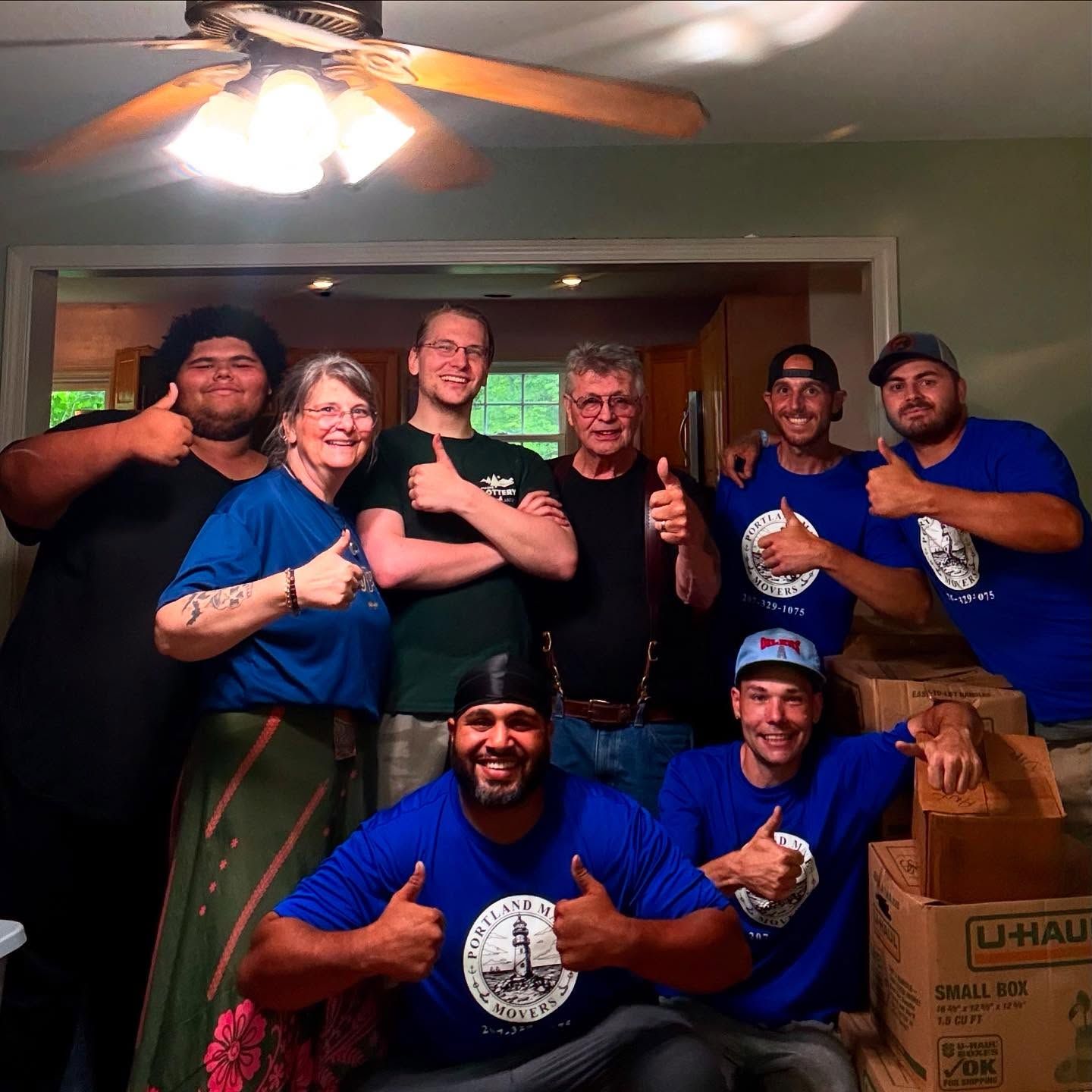 Group of people smiling, giving thumbs-up. Several wear blue shirts with a logo, boxes in background.