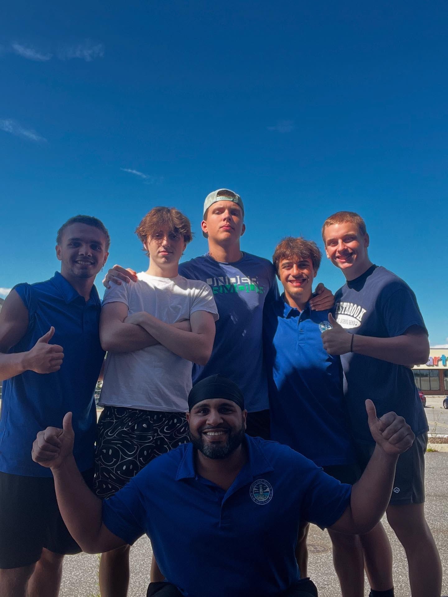 Six people in blue shirts pose with thumbs up against a blue sky, outside.