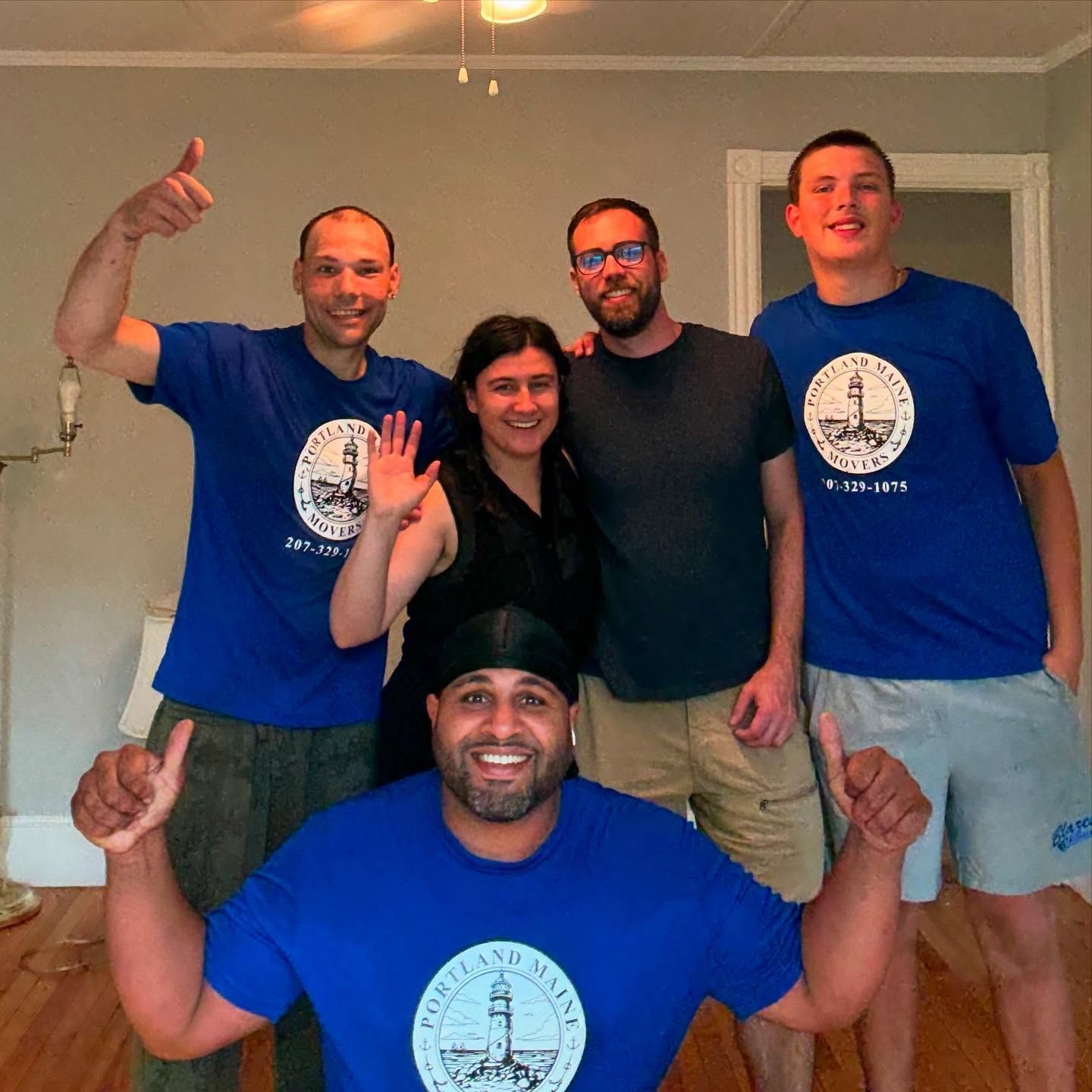 Five people, mostly wearing blue shirts, pose with thumbs up. One waves. Indoors, neutral walls.