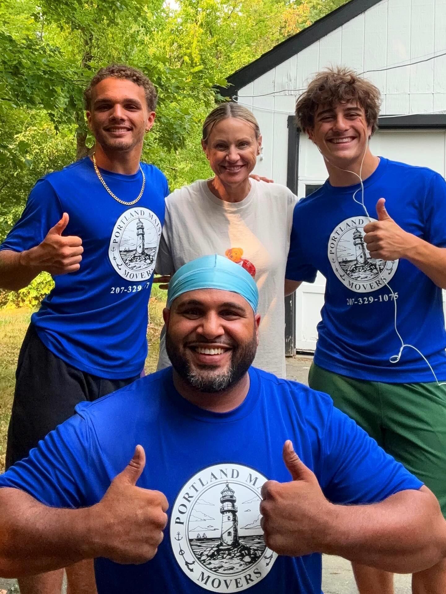 Four people smiling, thumbs up. Wearing blue shirts with a lighthouse logo, one with a swim cap. Standing outside a white building.