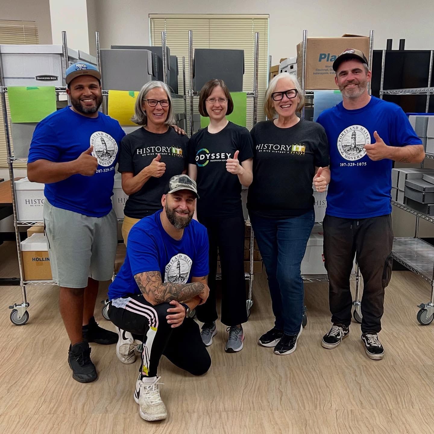 Group of six people smiling, giving thumbs up, wearing matching blue and black t-shirts in a storage space.