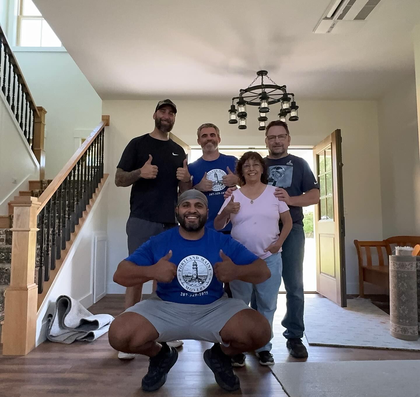 Group of people giving thumbs up in a home. One man squats, others stand near a staircase and front door.