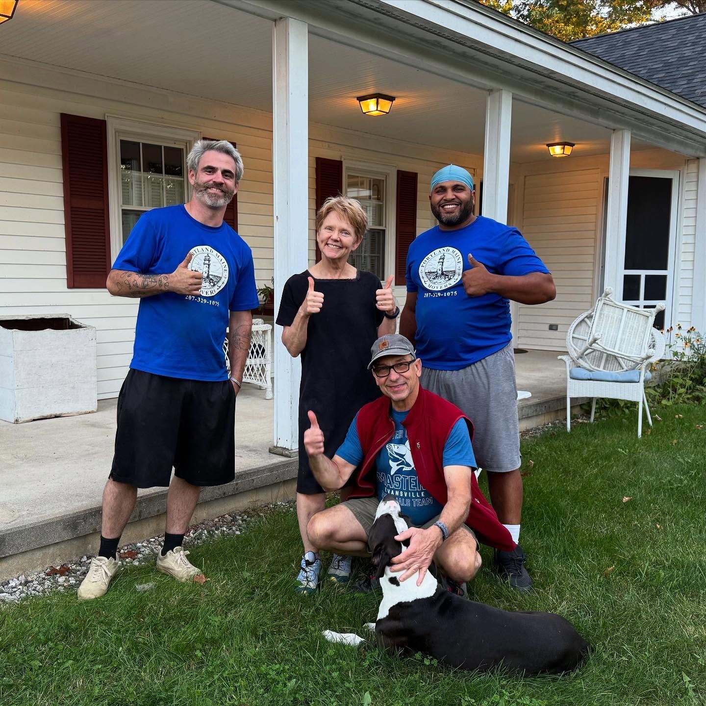 Four people giving thumbs up pose outside a house with a dog.