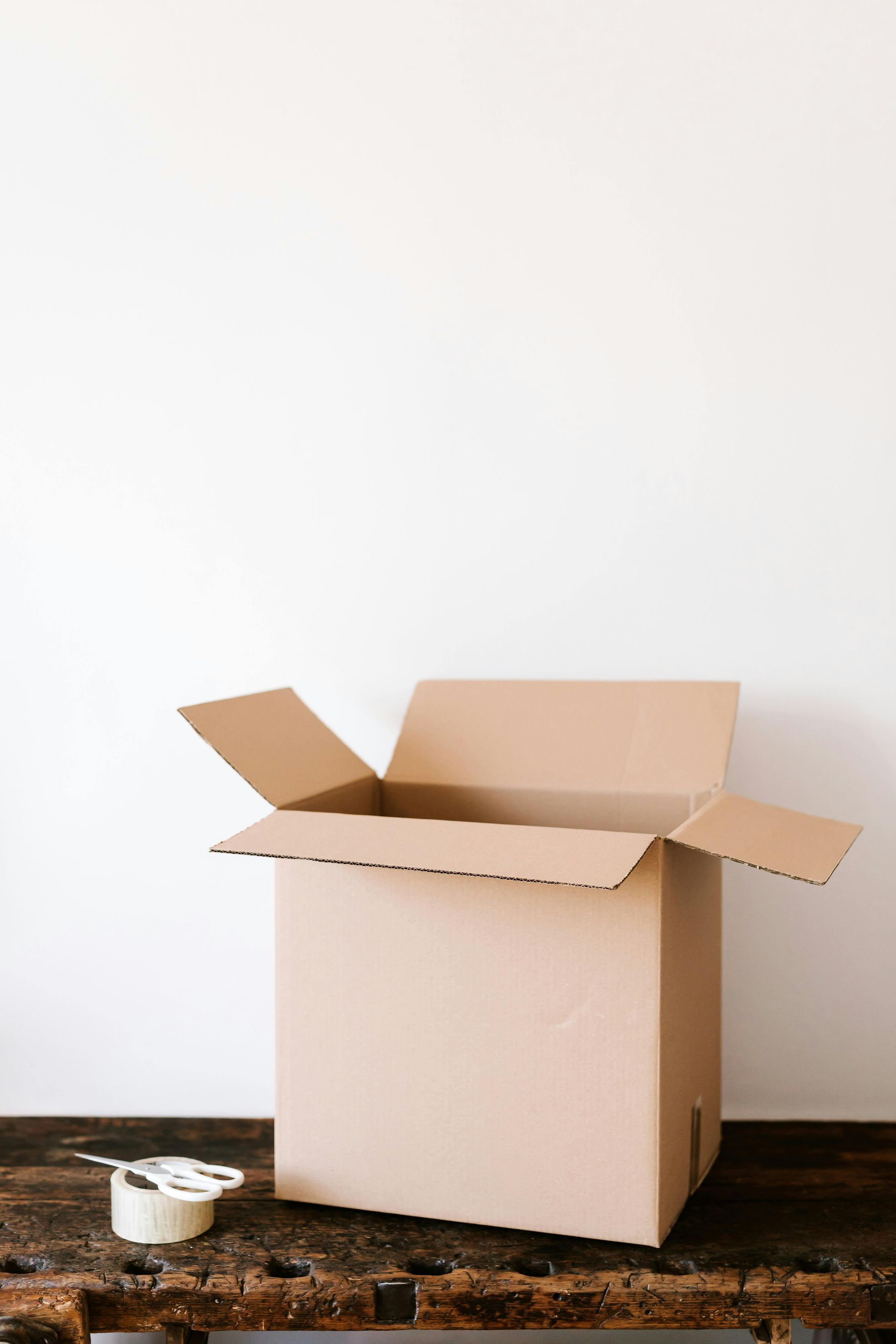 Open cardboard box on a rustic wooden surface with tape dispenser in front of a white wall.