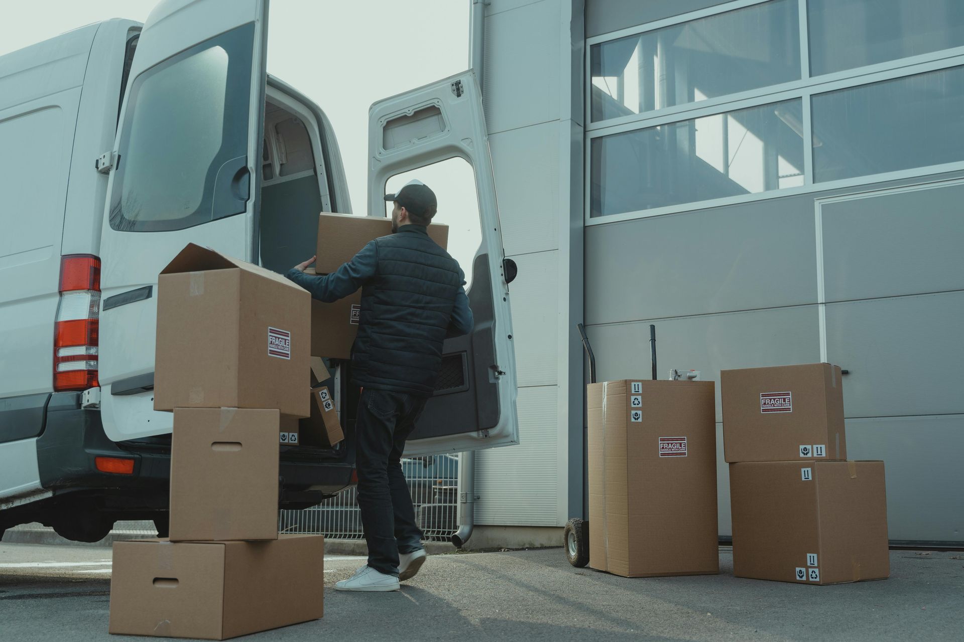 Man loading cardboard boxes into a white delivery van near a building with a large door.